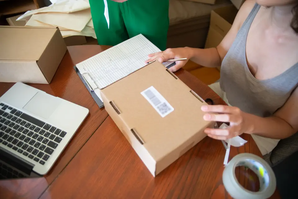 Hands packing an online order into a cardboard box beside a laptop and an order sheet