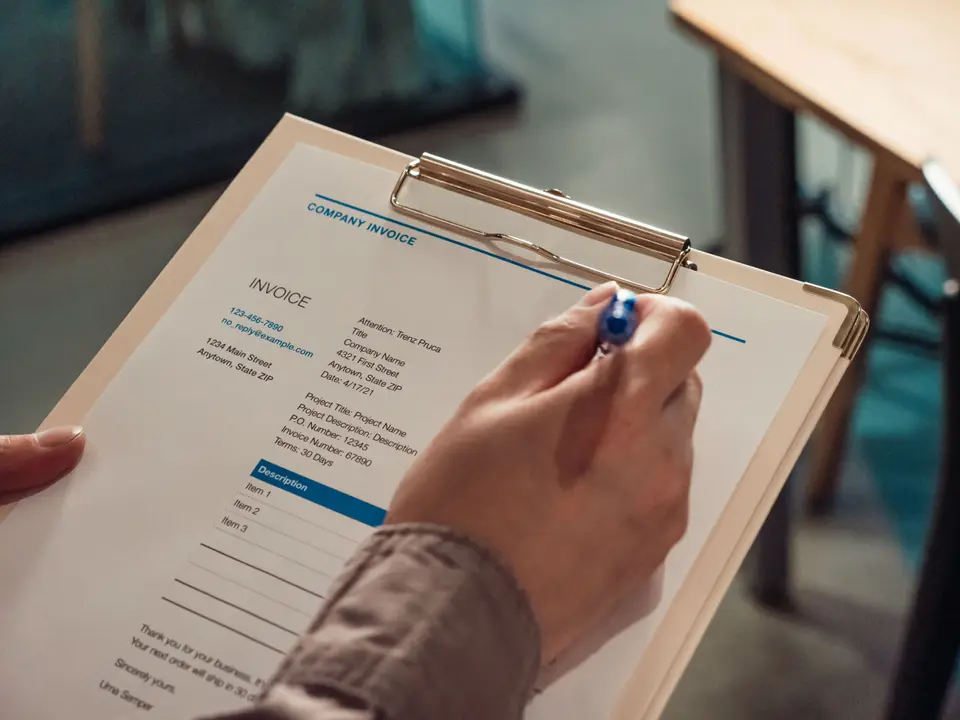 A hand filling in an invoice on a clipboard with a pen