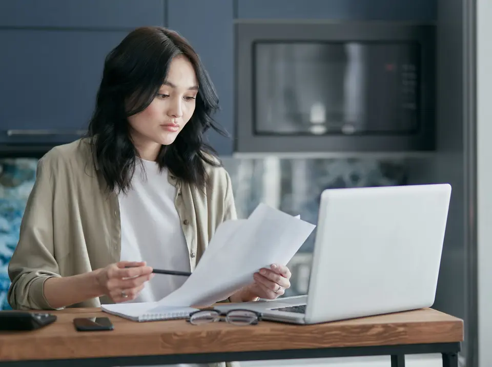 A bookkeeper reviewing printed documents beside a laptop at a home office desk
